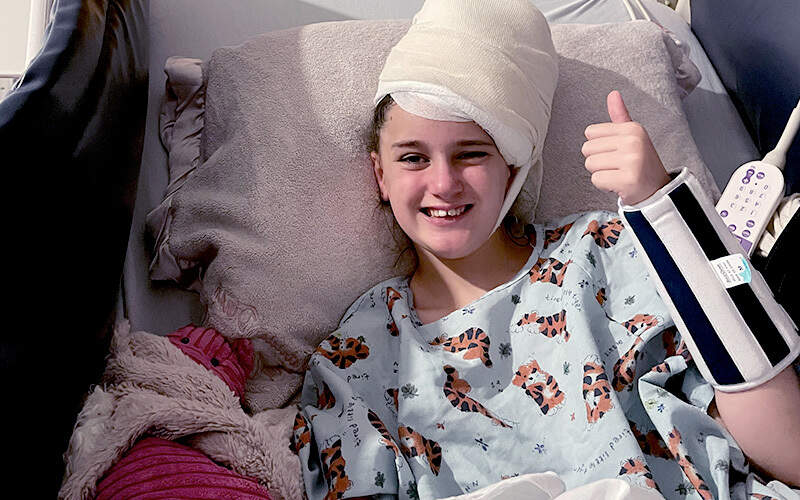 Young girl with bandaged head smiling and giving thumbs up from a hospital bed